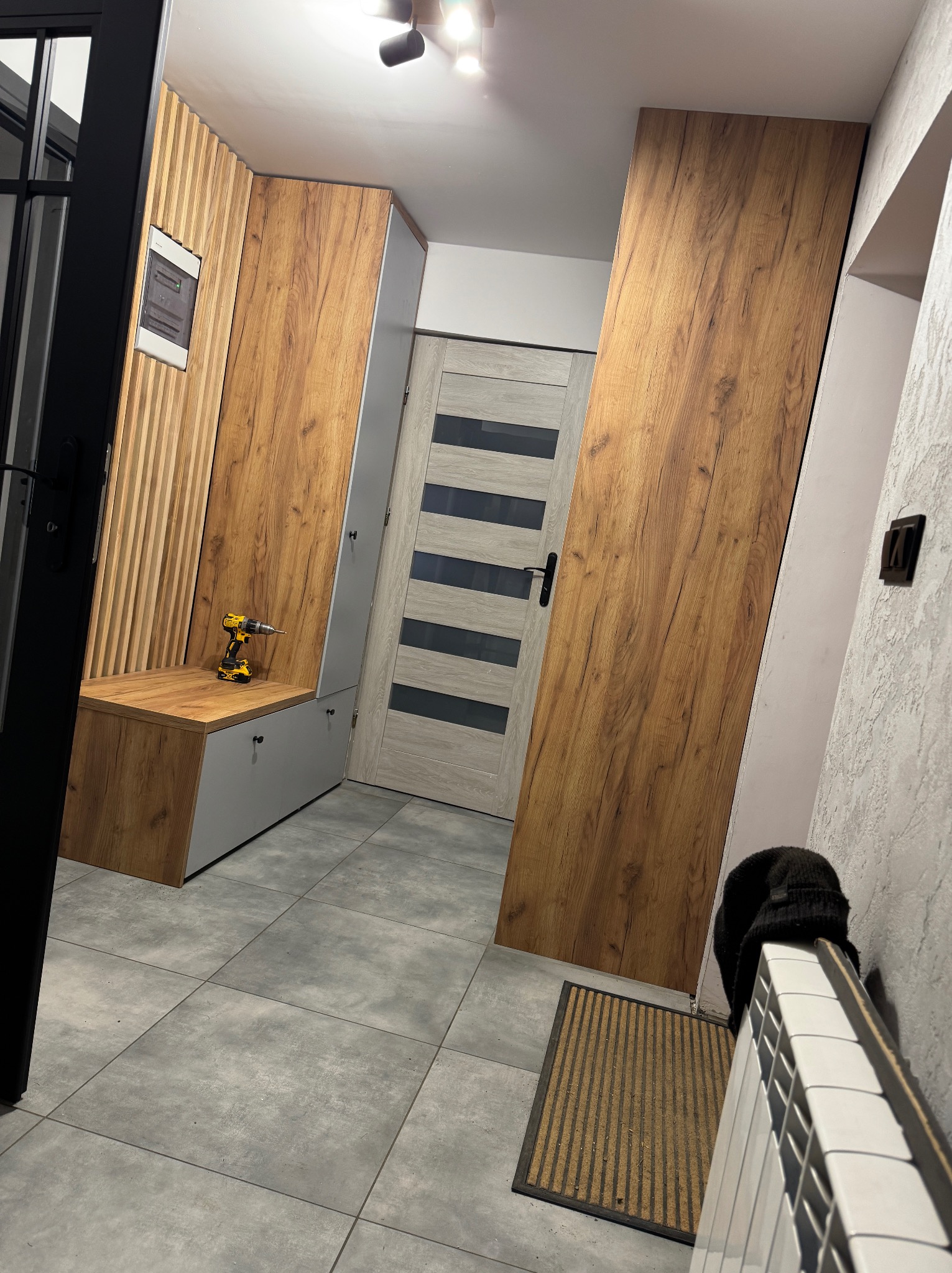 Drewniano-szara zabudowa przedpokoju z siedziskiem i szafkami, z wiertarką na blacie. Widoczna podłoga z szarych płytek i drzwi wejściowe.
