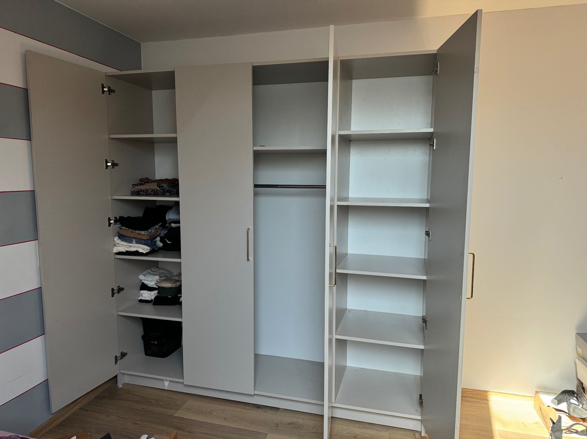 Jasnoszara szafa trzydrzwiowa z otwartymi drzwiami, ukazująca półki z ubraniami i drążek na wieszaki. Minimalistyczny design, widoczne okucia i drewniana podłoga.