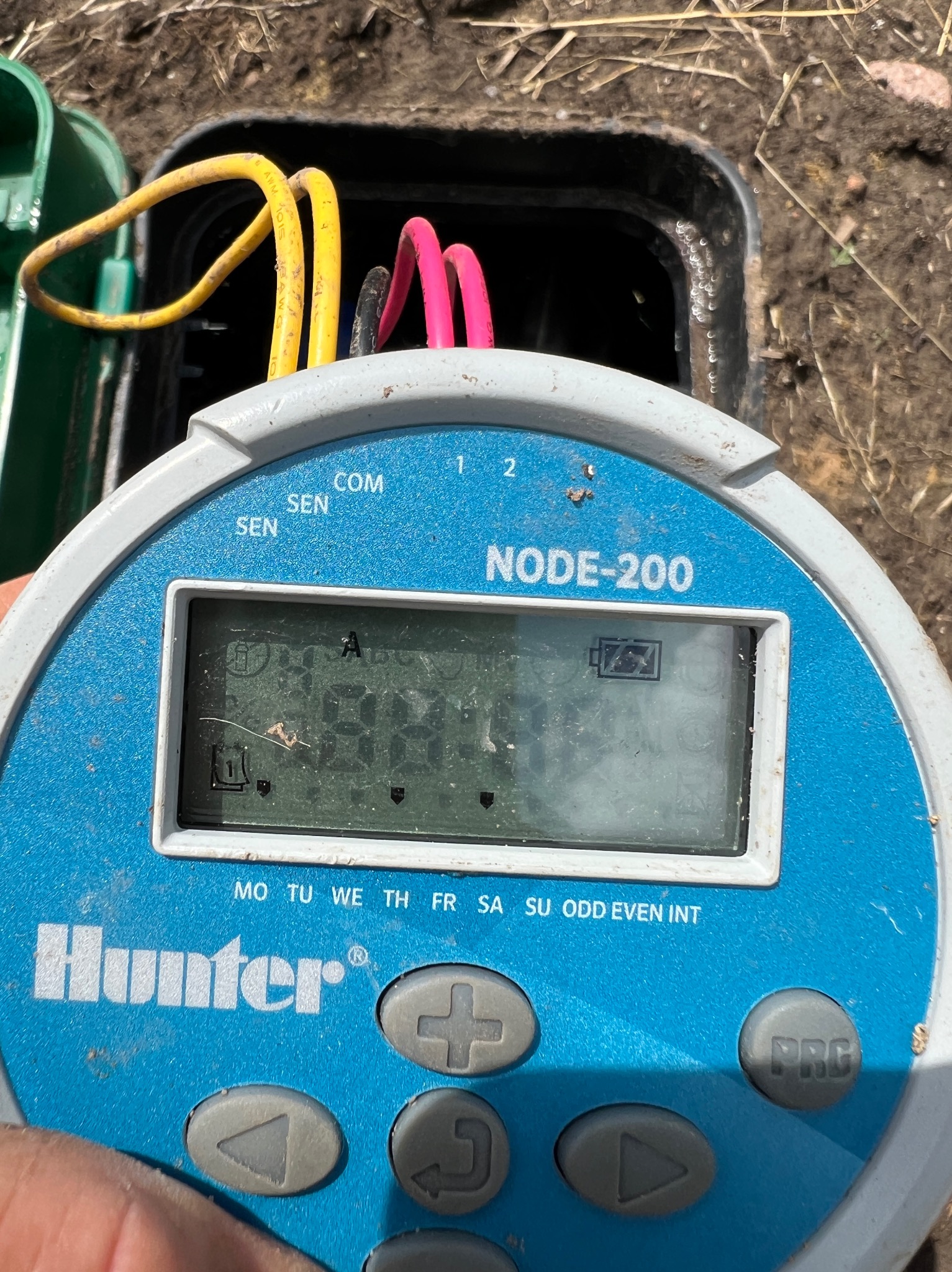 Programator Hunter NODE-200 do systemów nawadniania, widoczne przewody i otwarty zawór. Kontroler z wyświetlaczem i przyciskami, zasilany bateryjnie.