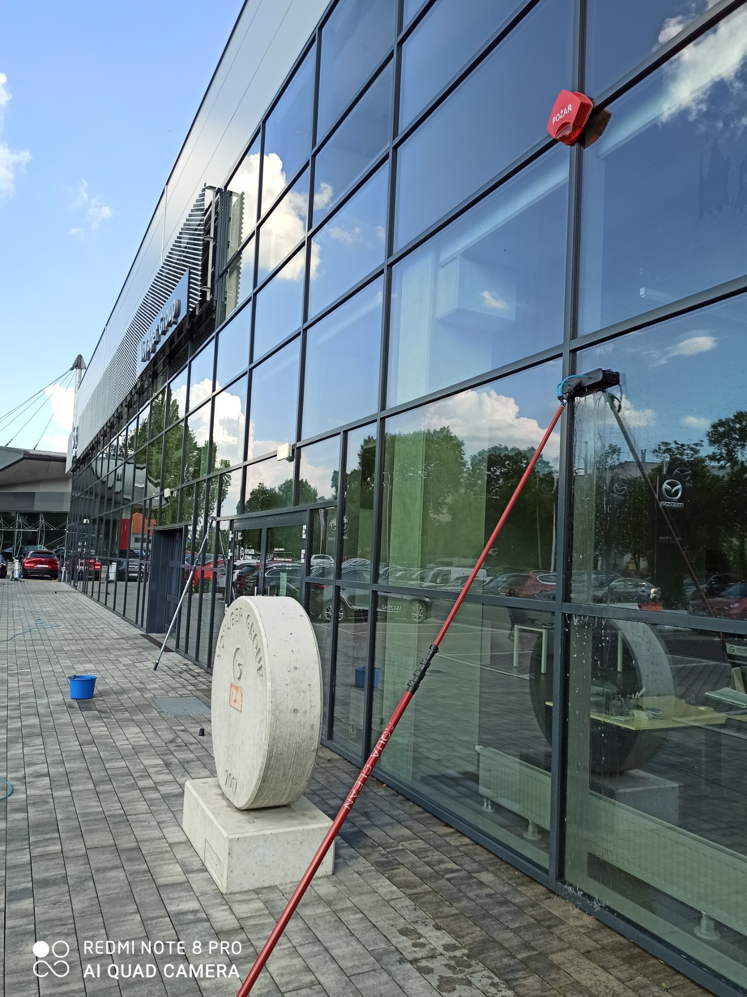 Mycie okien budynku z użyciem lancy teleskopowej. Widoczne odbicia w czyszczonych szybach, element dekoracyjny przed budynkiem i sprzęt do mycia okien.