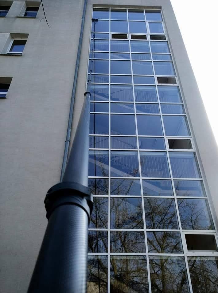 Elewacja budynku z wieloma oknami, czyszczona teleskopową lancą. W oknach odbija się drzewo. Widok z dołu, lanca na pierwszym planie.