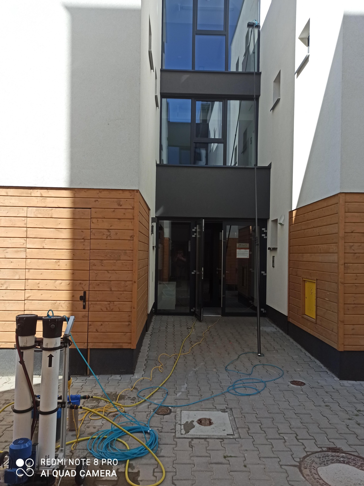 Urządzenie do mycia okien ciśnieniowego z lancą teleskopową przed budynkiem z przeszkloną fasadą. Widoczne przewody doprowadzające wodę.