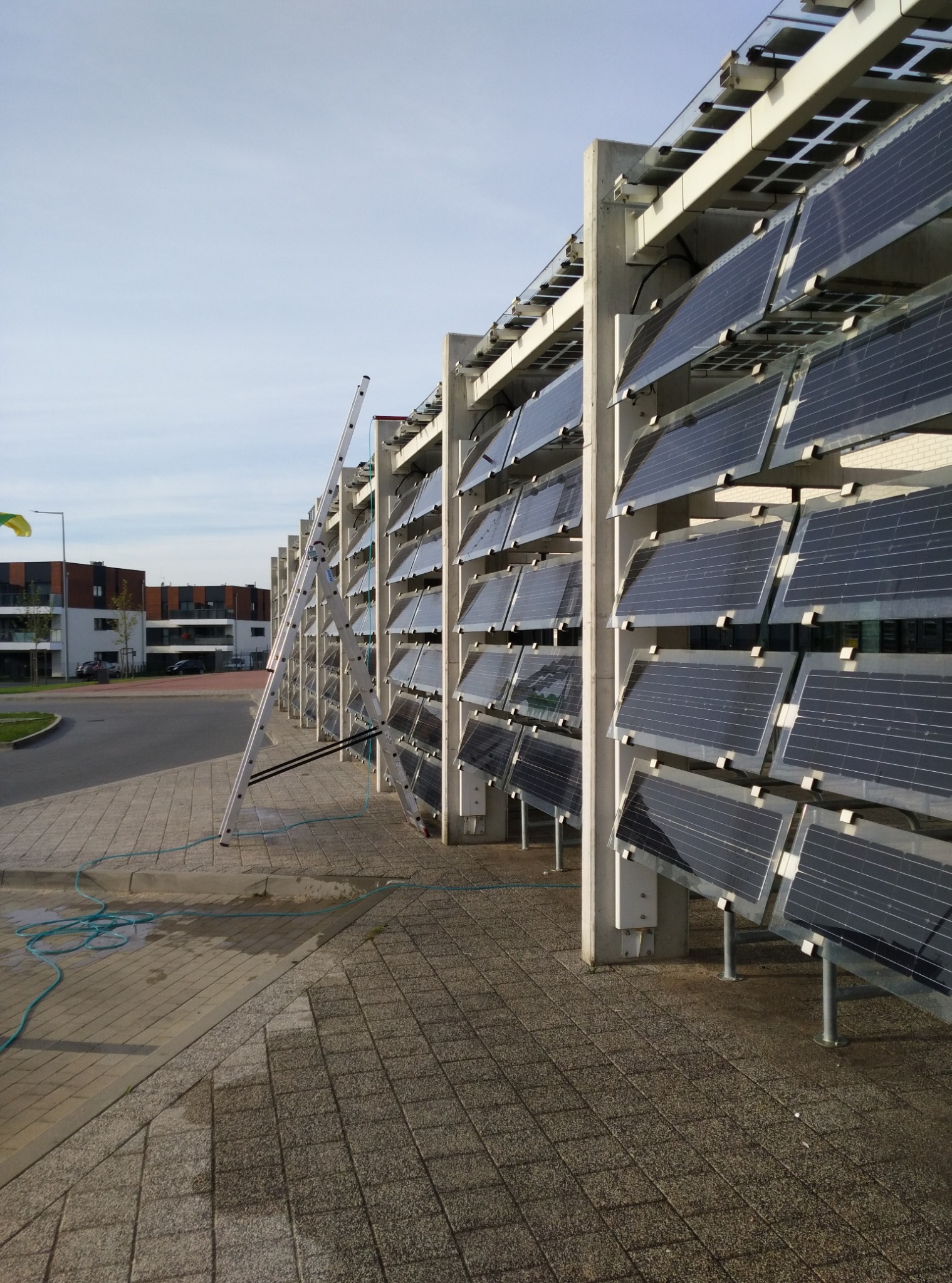 Mycie paneli słonecznych na konstrukcji wsporczej. Rozstawiona drabina, widoczny wąż doprowadzający wodę. Tło: budynki mieszkalne, chodnik z kostki.