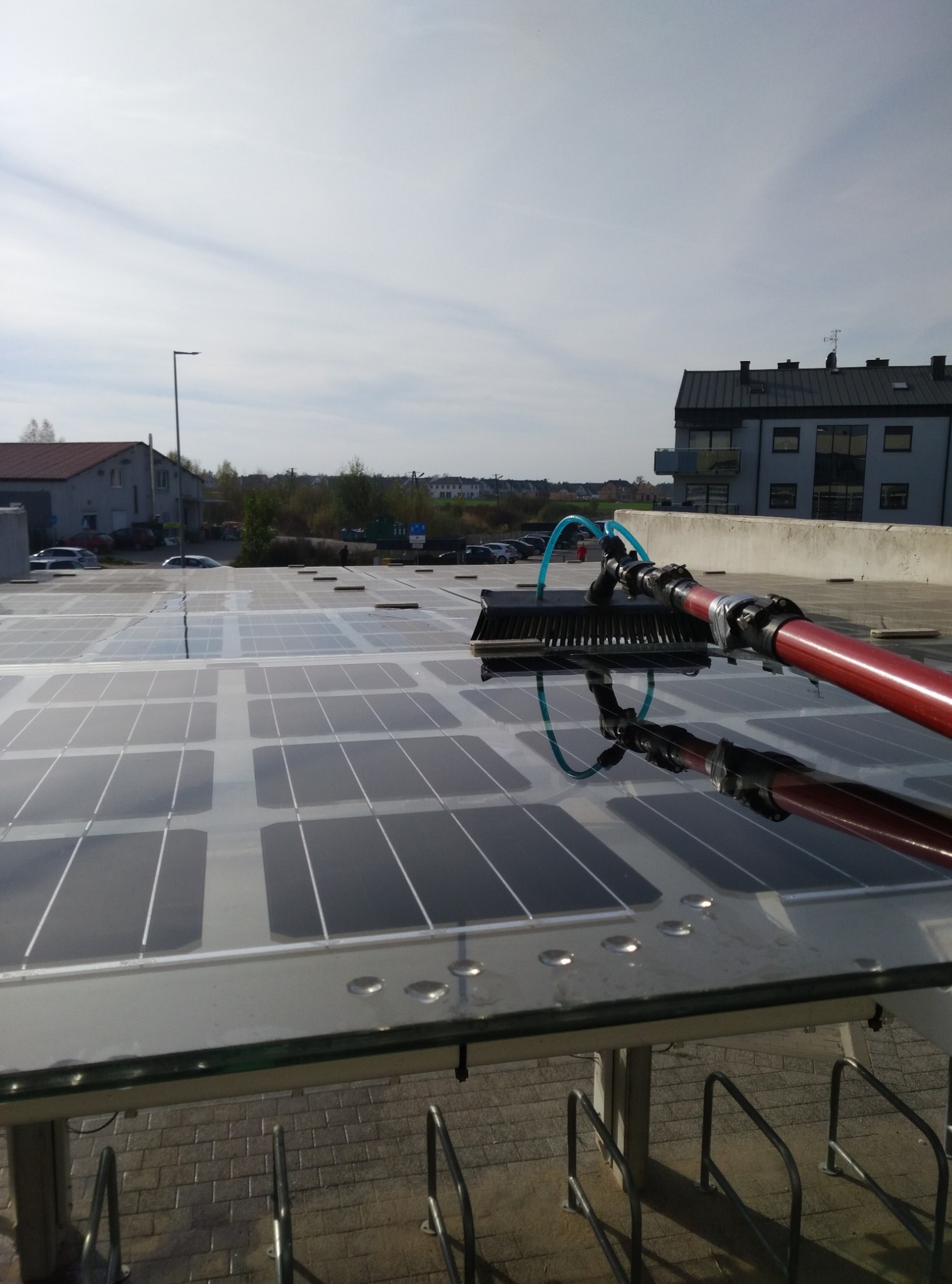 Czyszczenie paneli słonecznych na dachu budynku za pomocą szczotki na długim wysięgniku, w tle widoczne budynki i błękitne niebo.