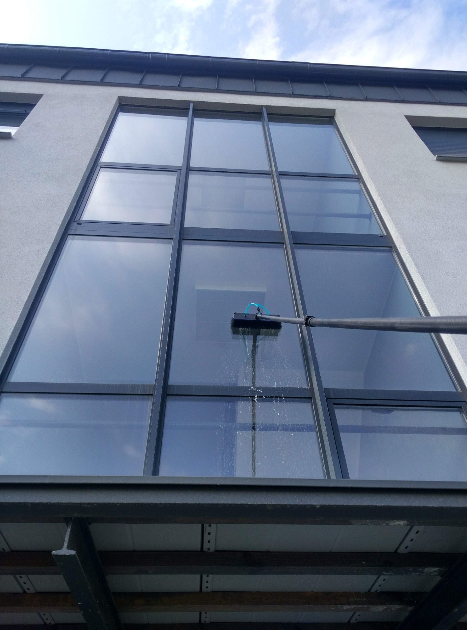 Czyszczenie dużych okien na wysokości w Błażejewie za pomocą szczotki na teleskopowym kiju. Woda spływa po szybie. Widok z dołu na budynek.