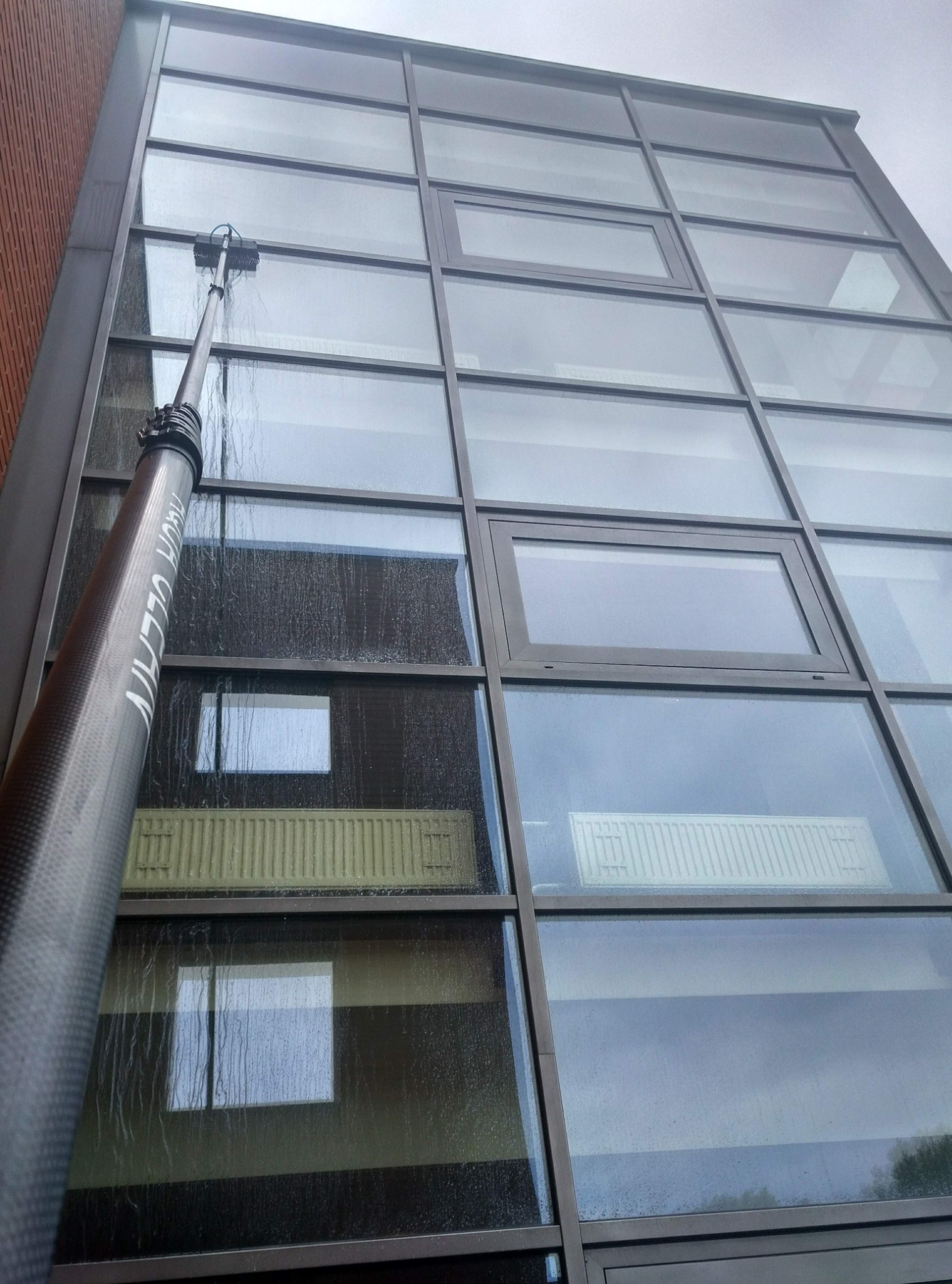 Mycie okien na wysokości w budynku biurowym za pomocą teleskopowej lancy. Widoczne smugi na szybach i odbicie otoczenia w czyszczonych oknach.