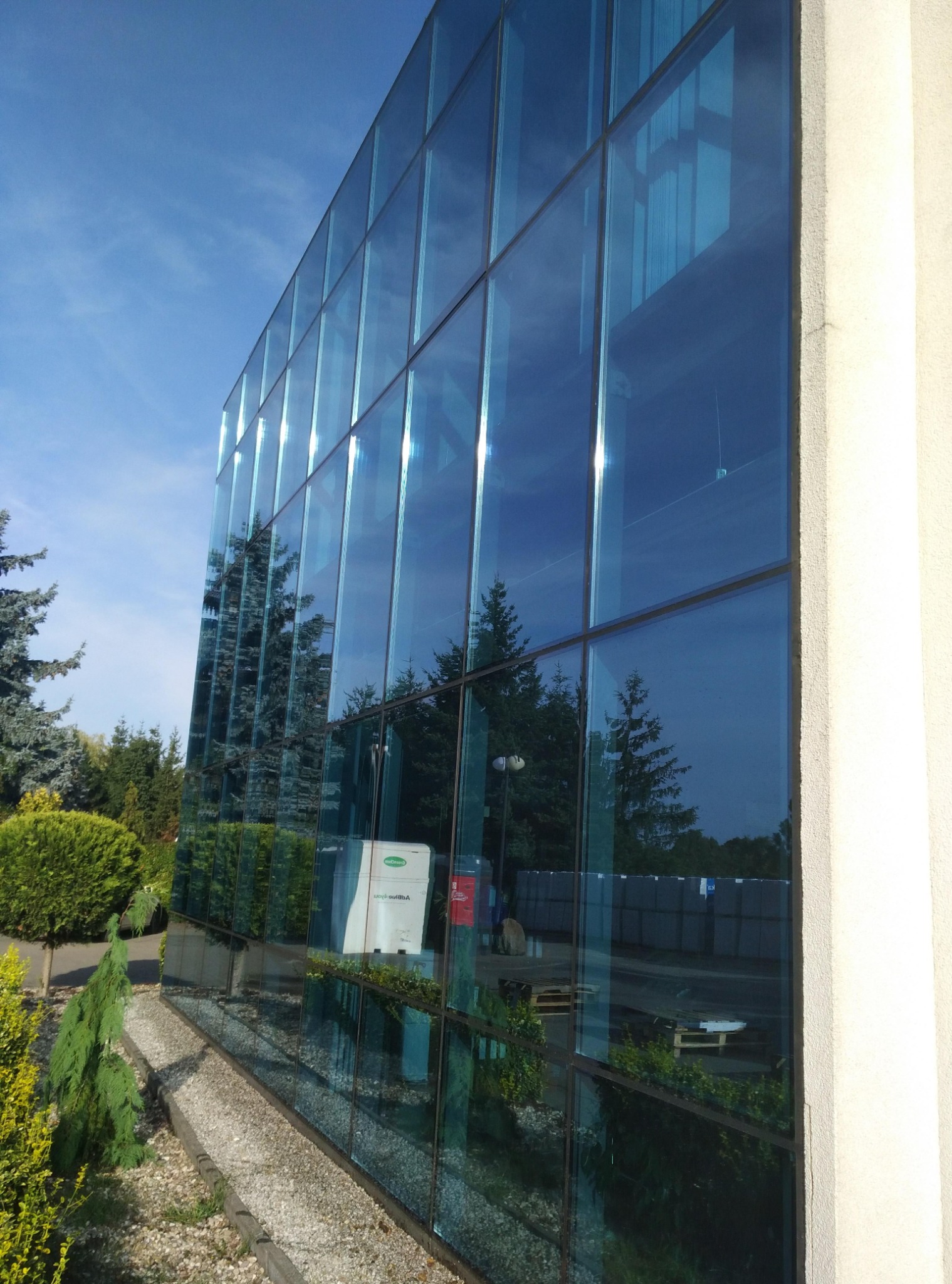 Szklana fasada budynku biurowego odbija błękitne niebo i zieleń drzew. Widoczne detale architektoniczne i otoczenie budynku. Perspektywa z dołu, podkreślająca wysokość.