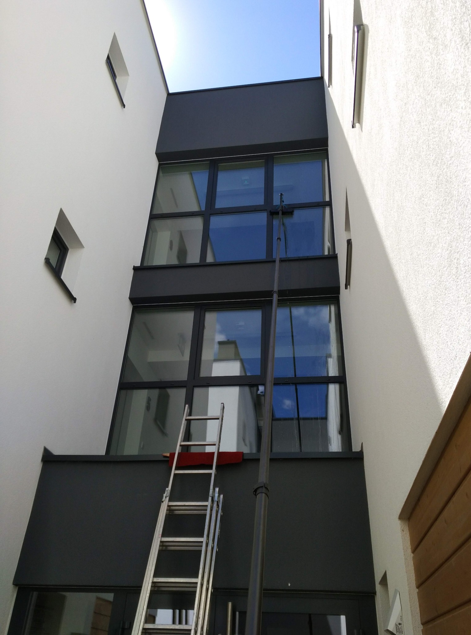 Czyszczenie okien na wysokości w nowoczesnym budynku z użyciem drabiny i tyczki teleskopowej. Widok z dołu, błękitne niebo w tle.