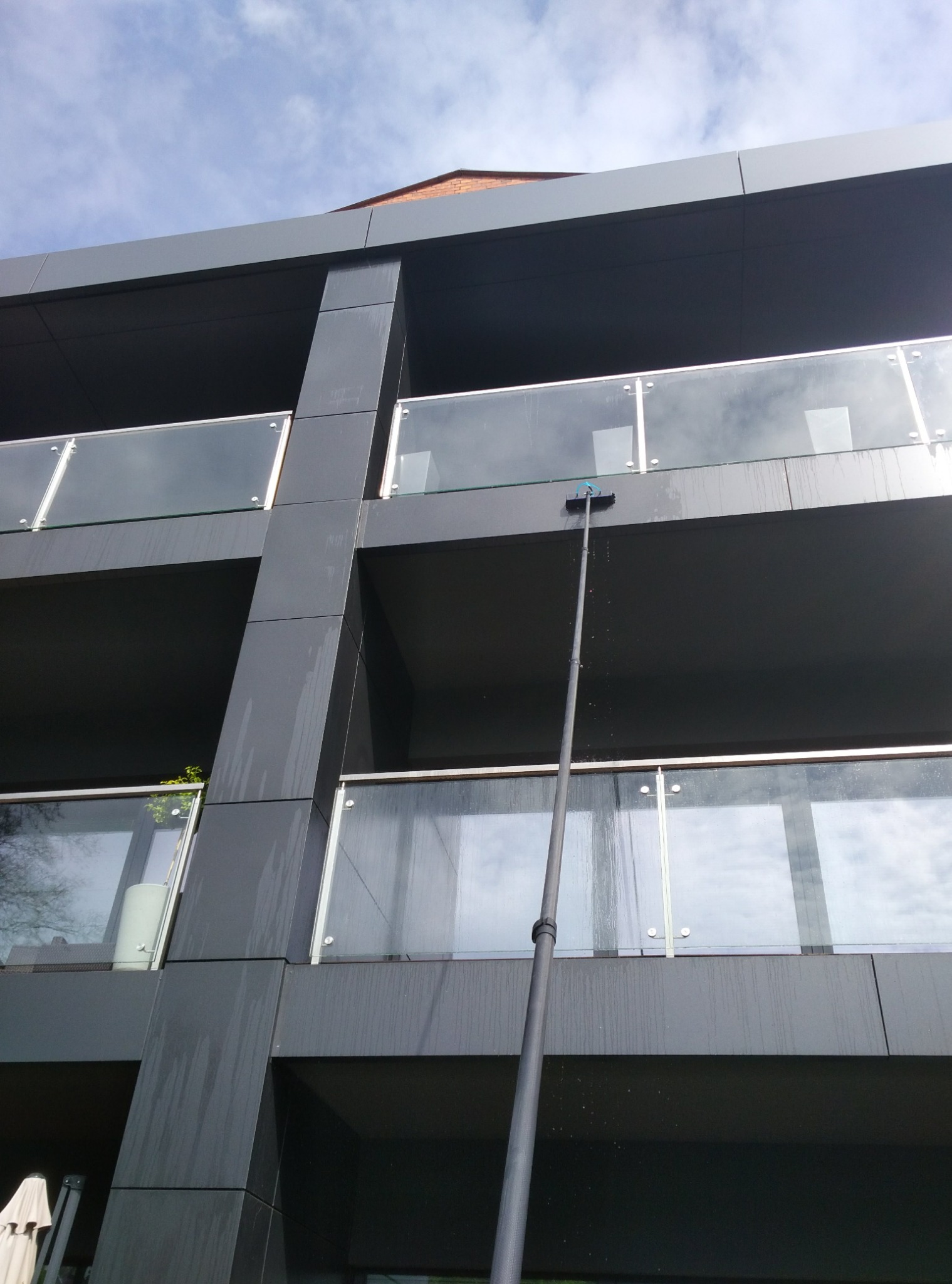 Mycie okien na wysokości nowoczesnego budynku z ciemną elewacją. Długa tyczka z szczotką sięga do okien na balkonie. Widoczne ślady wody na elewacji.