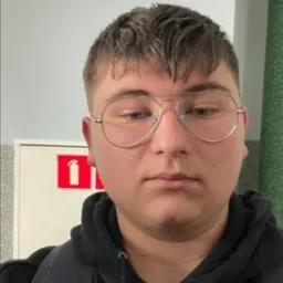 Portret młodego mężczyzny w okularach typu aviator, ubrany w czarną bluzę z kapturem, z plecakiem na ramieniu, na tle ściany i skrzynki z gaśnicą.