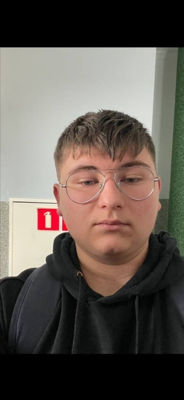 Portret młodego mężczyzny w okularach typu aviator, ubrany w czarną bluzę z kapturem, z plecakiem na ramieniu, na tle ściany i skrzynki z gaśnicą.