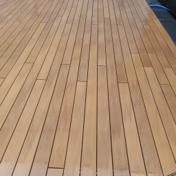 Grzegorz Karabin Czysta Przestrzeń - Wyczyszczona, mokra drewniana podłoga tarasu z widocznymi smugami wody i fragmentem niebieskiego węża w rogu, sugerująca świeżo zakończone mycie.