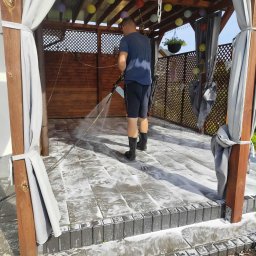 Grzegorz Karabin Czysta Przestrzeń - Czyszczenie kostki brukowej na tarasie altany z dekoracjami. Mężczyzna w gumowcach używa myjki ciśnieniowej, spłukując brud z szarej kostki. Widoczne drewniane elementy konstrukcji.