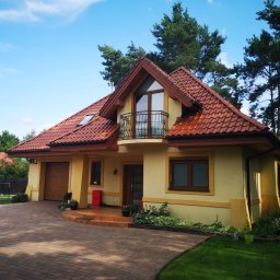 Grzegorz Karabin Czysta Przestrzeń - Ukończony dom jednorodzinny z balkonem, z czerwoną dachówką i żółtą elewacją, otoczony zielenią i kostką brukową. Widok z przodu, słoneczny dzień.