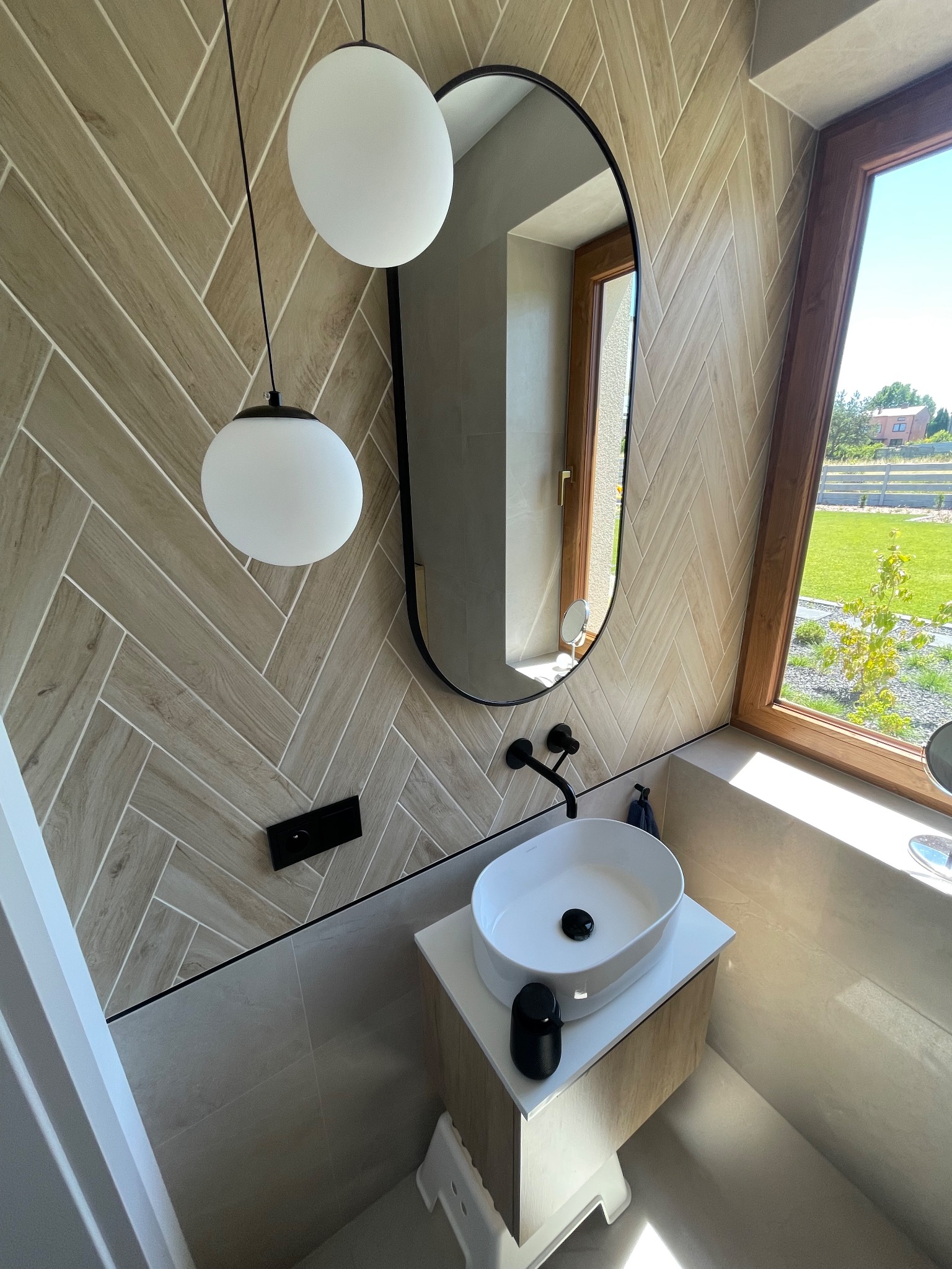 Nowoczesna łazienka z umywalką nablatową, lustrem w czarnej ramie i heksagonalną okładziną ścienną w odcieniach beżu, oświetlona wiszącymi lampami.