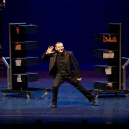 Uśmiechnięty iluzjonista prezentuje trik z asystentkami w skrzyniach na kółkach podczas występu na scenie z niebieskim tłem i oświetleniem.