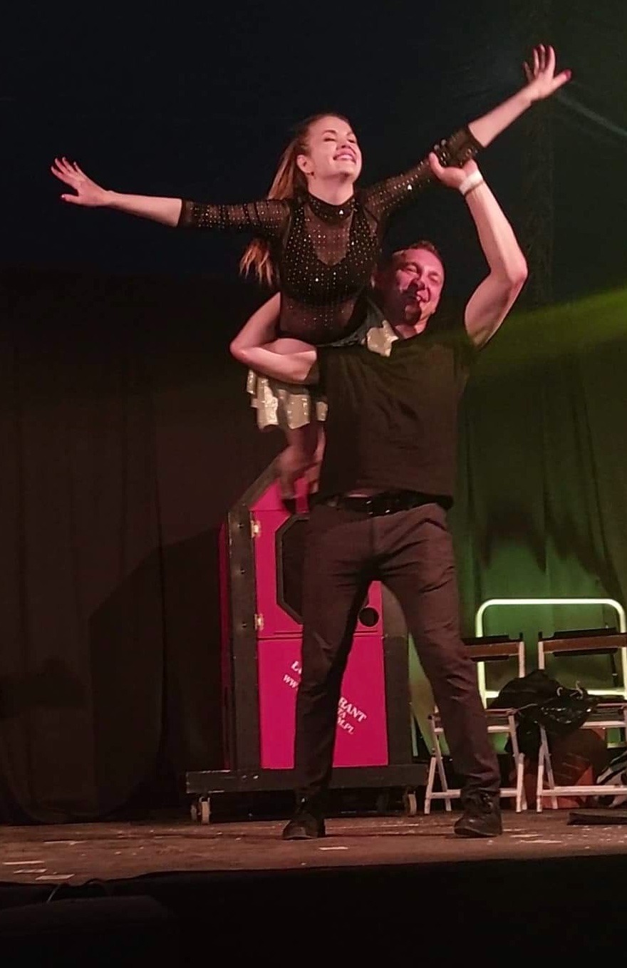 Iluzjonista podnosi uśmiechniętą asystentkę w błyszczącej sukience podczas występu na scenie z rekwizytami. Czarny strój iluzjonisty.