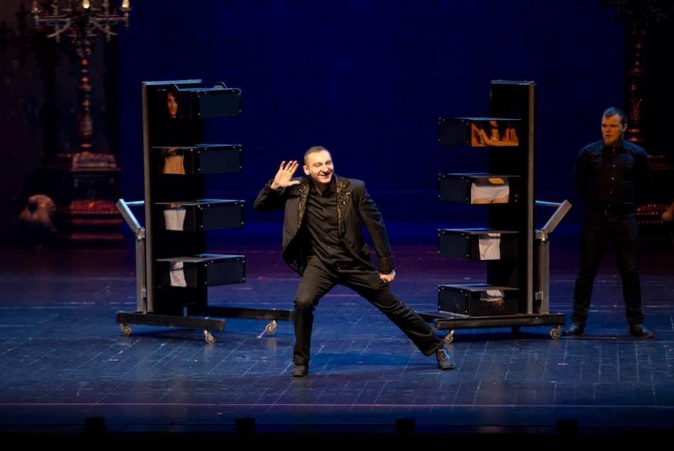 Uśmiechnięty iluzjonista prezentuje trik z asystentkami w skrzyniach na kółkach podczas występu na scenie z niebieskim tłem i oświetleniem.