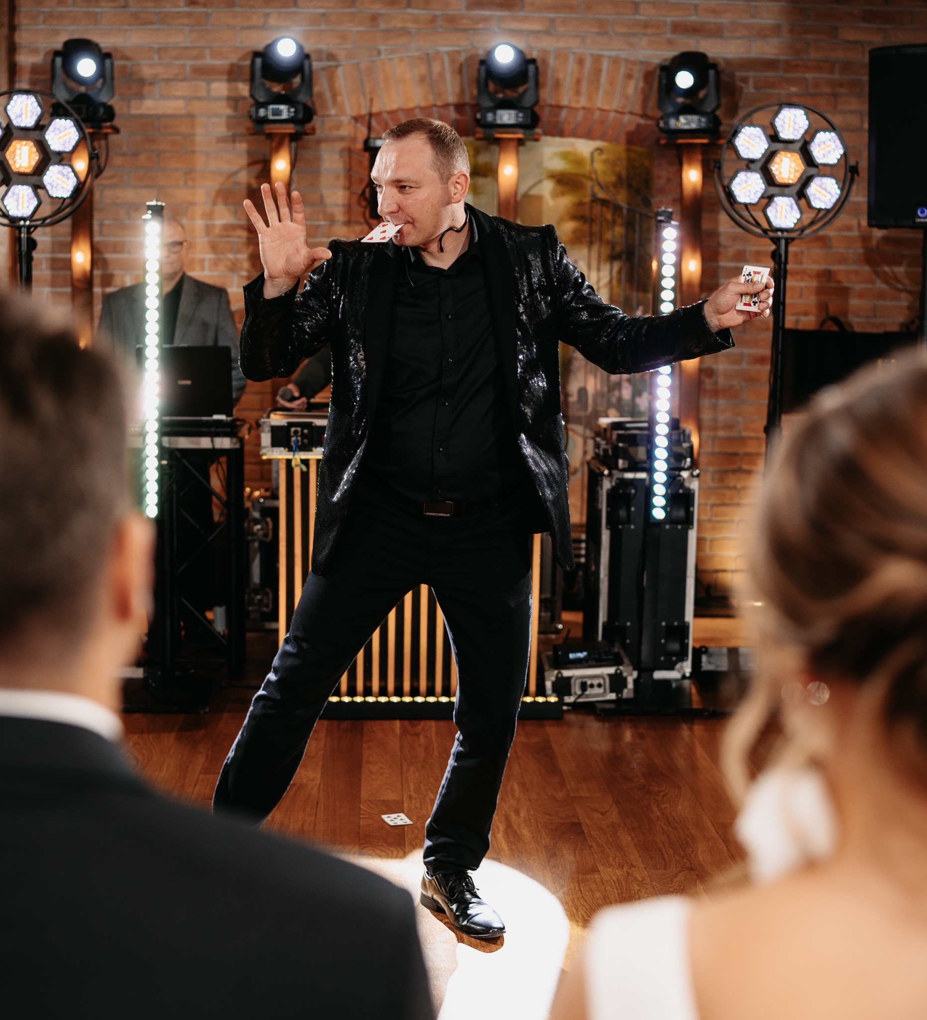 Iluzjonista w cekinowej marynarce prezentuje trik z kartami na scenie z oświetleniem, widzowie obserwują z bliska. Karty unoszą się w powietrzu.