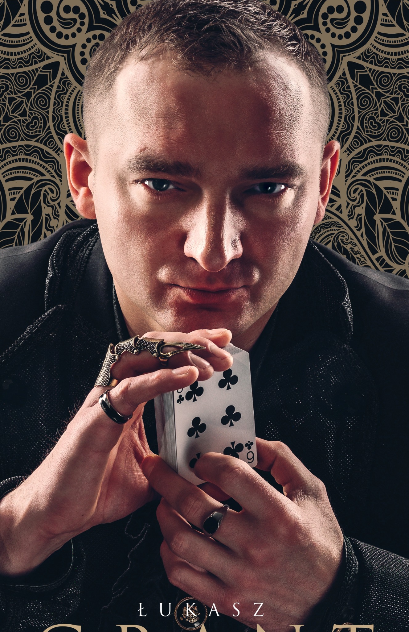 Portret iluzjonisty Łukasza Grant z kartami w dłoniach, na tle wzorzystej tapety. Uwagę przykuwają pierścienie na palcach.