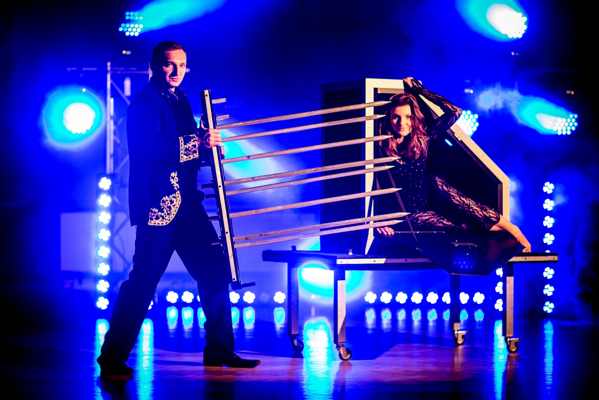 Iluzjonista w zdobionej marynarce prezentuje trik z przecinaniem kobiety w skrzyni za pomocą ostrzy na tle niebieskich świateł scenicznych.