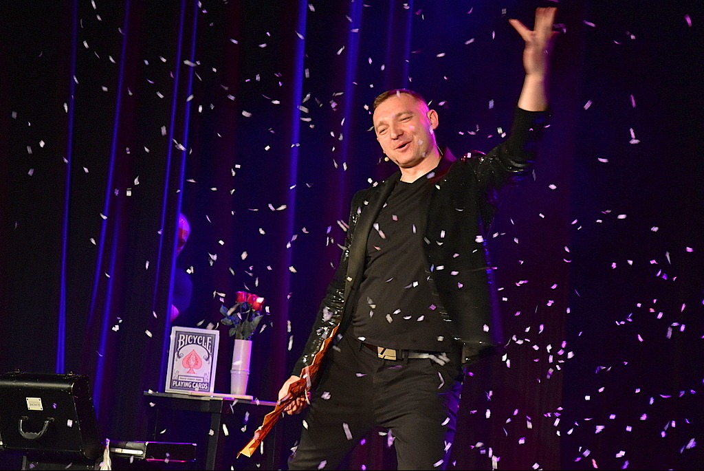 Iluzjonista w czarnym cekinowym żakiecie gestykuluje na scenie w deszczu konfetti. W tle zasłony, stolik z kartami Bicycle i różami w wazonie.