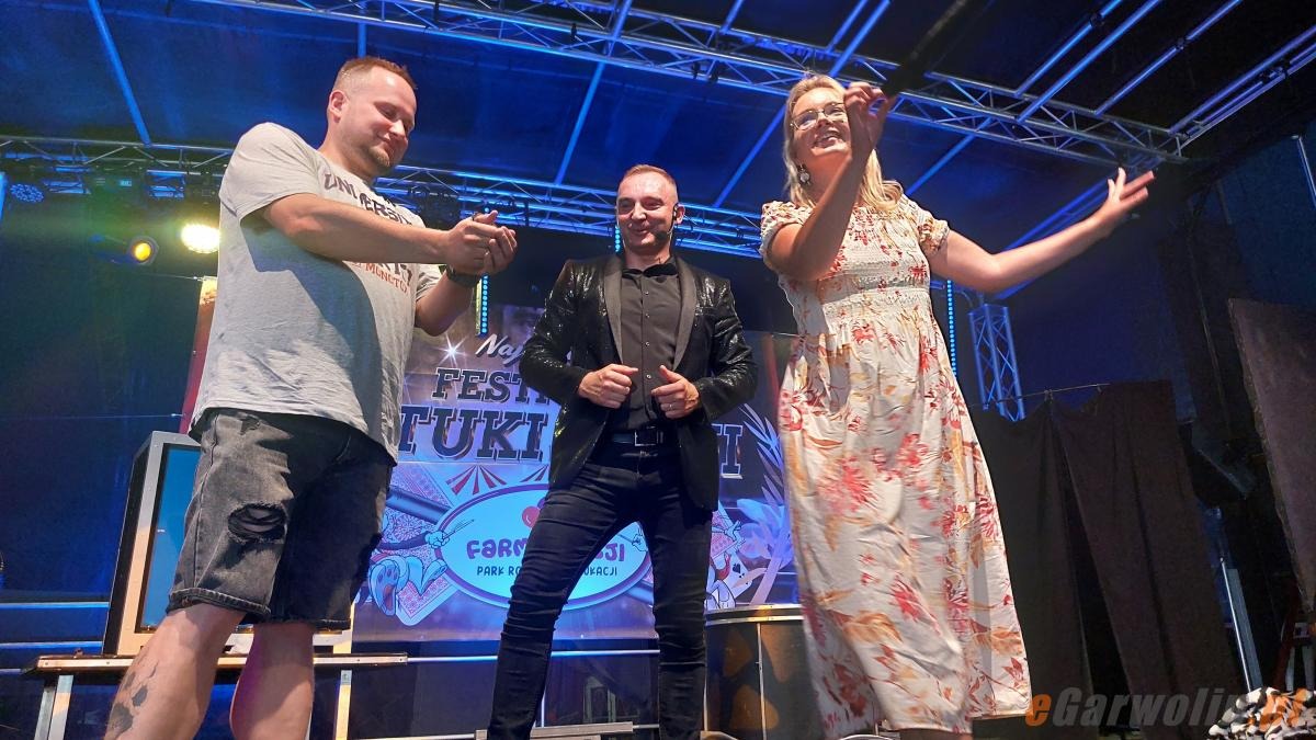 Iluzjonista w błyszczącej marynarce na scenie z publicznością podczas festiwalu. W tle baner z nazwą wydarzenia.