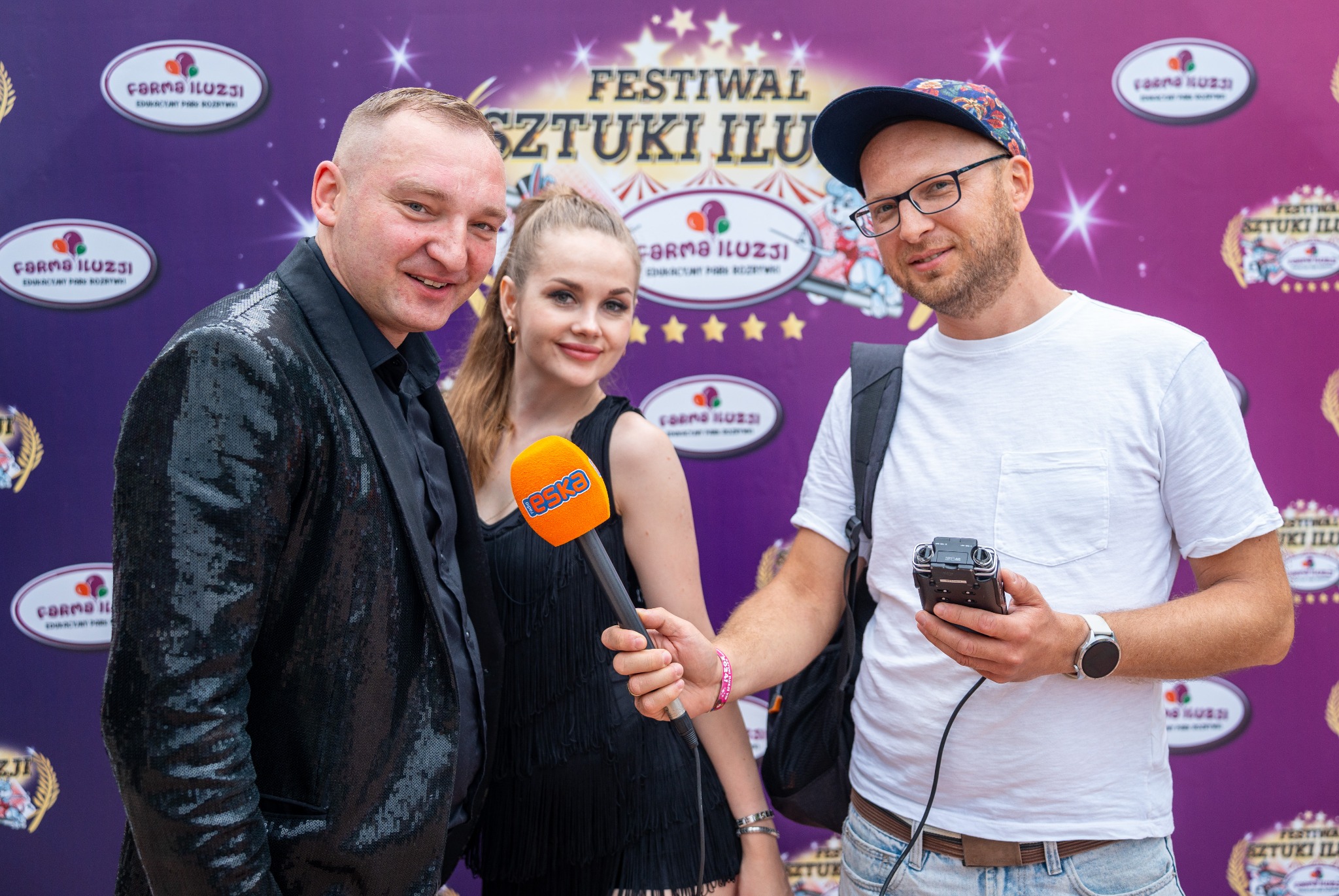 Trzy osoby na tle fioletowej ścianki z logo Festiwalu Sztuki Iluzji. Mężczyzna w cekinowej marynarce, kobieta z mikrofonem i mężczyzna z rejestratorem.