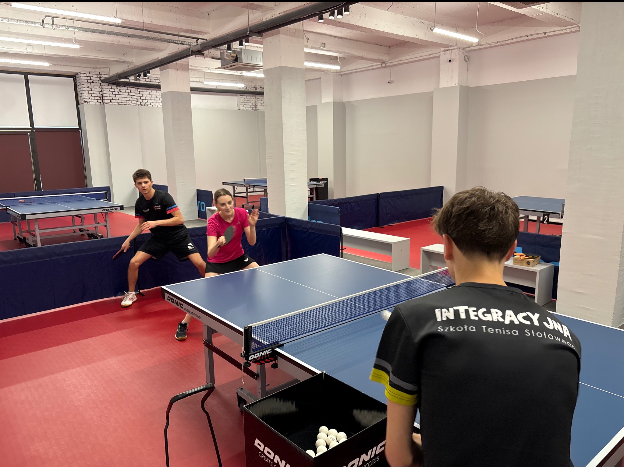 Trening tenisa stołowego w hali: dwoje graczy w akcji, trener obserwuje. Logo szkoły tenisa na koszulce trenera. Profesjonalne stoły Donic.