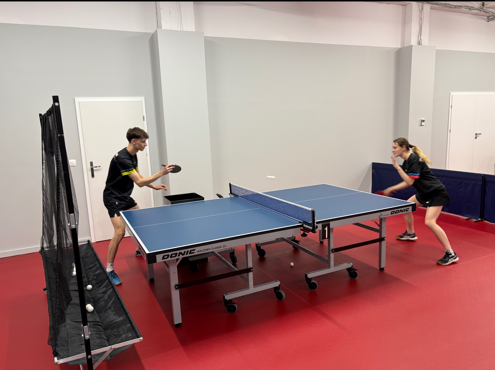 Sala do tenisa stołowego z dwoma graczami w akcji, widoczny stół marki Donic, siatka do zbierania piłek i czerwona podłoga sportowa. Jasne oświetlenie.