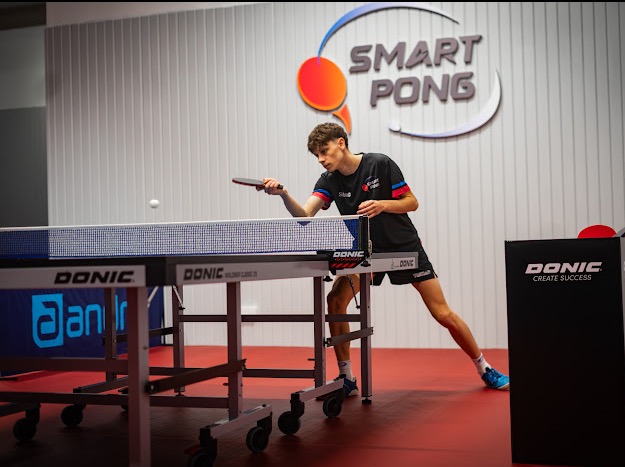 Młody mężczyzna serwuje piłeczkę do tenisa stołowego przy stole marki Donic w jasnym pomieszczeniu z logo Smart Pong na ścianie.