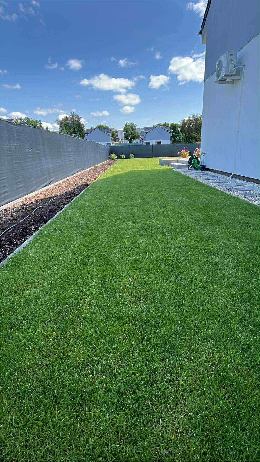 Zadbany trawnik z systemem nawadniania obok ogrodzenia i domu w słoneczny dzień. Krzewy ozdobne i kostka brukowa dopełniają estetyki ogrodu.