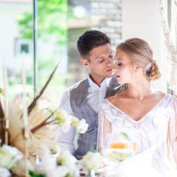 Wioletta Pietrucha Fotografia - Elegancka para młoda w jasnym wnętrzu, on w szarej kamizelce, ona w białej sukni z frędzlami, delikatne spojrzenie, dekoracje kwiatowe i makrama w tle.