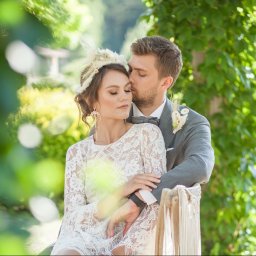 Wioletta Pietrucha Fotografia - Elegancka para młoda w plenerze: on w szarym garniturze, ona w koronkowej sukience i ozdobie z piór. Romantyczny uścisk na tle zieleni.