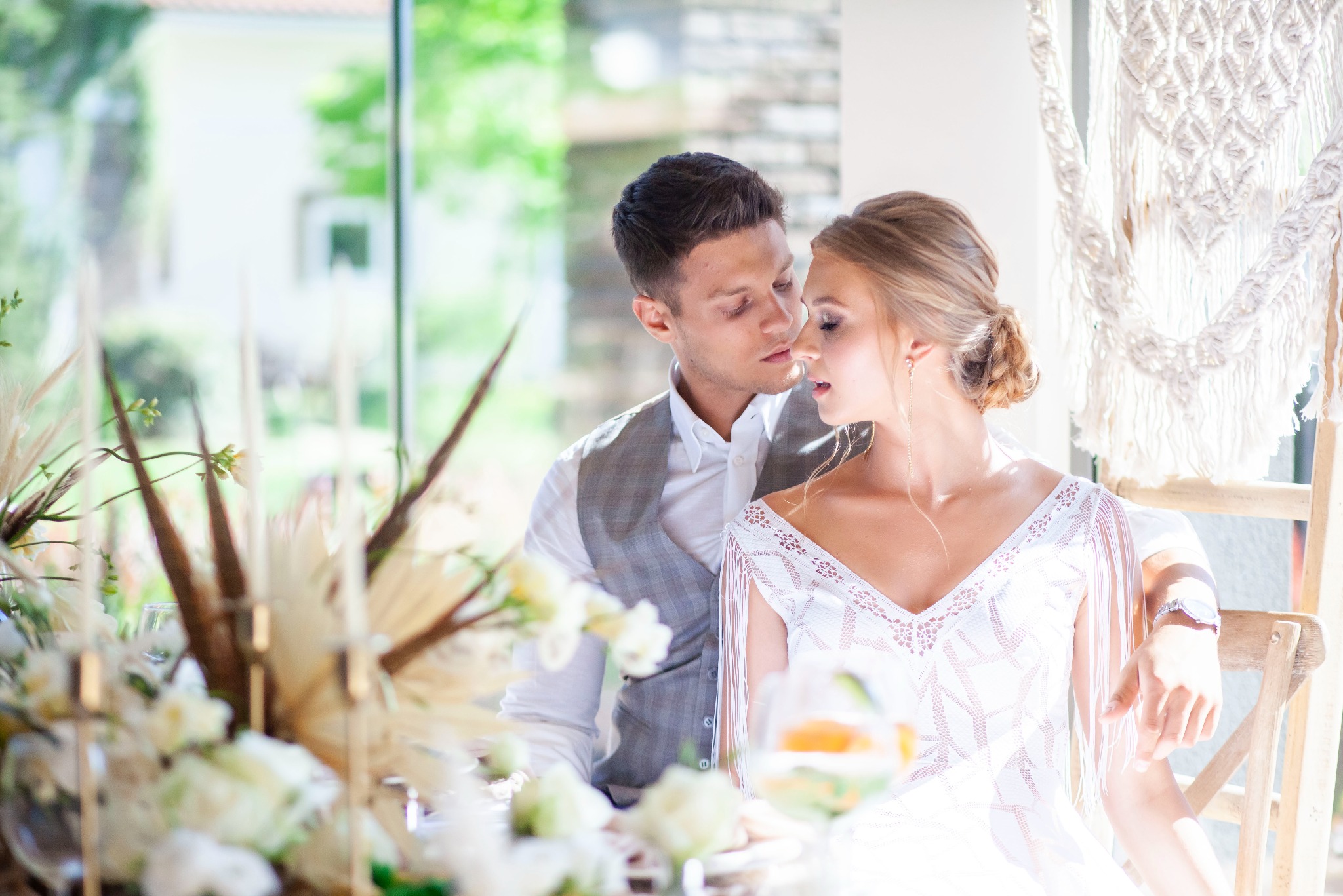 Elegancka para młoda w jasnym wnętrzu, on w szarej kamizelce, ona w białej sukni z frędzlami, delikatne spojrzenie, dekoracje kwiatowe i makrama w tle.