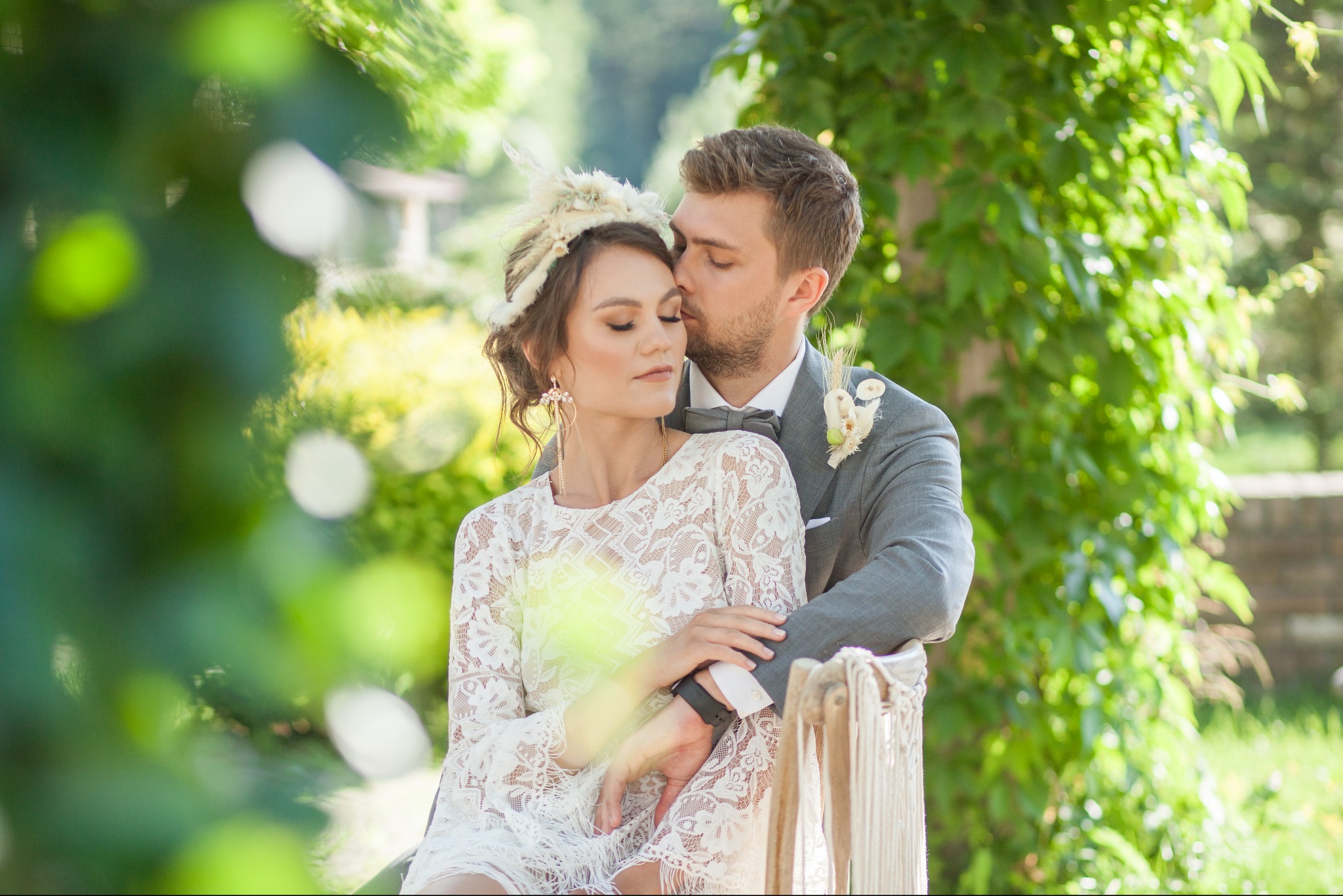 Elegancka para młoda w plenerze: on w szarym garniturze, ona w koronkowej sukience i ozdobie z piór. Romantyczny uścisk na tle zieleni.