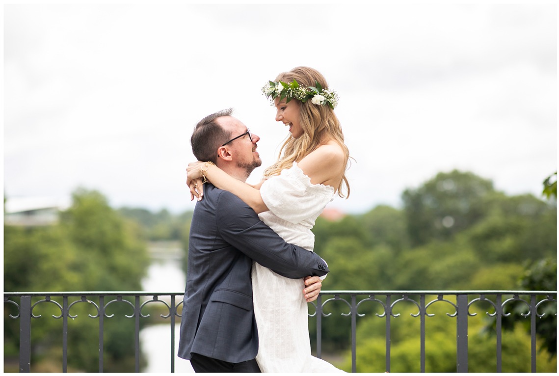 Uśmiechnięta panna młoda w wianku kwiatowym, trzymana w objęciach przez pana młodego w garniturze na tle zieleni i balustrady.