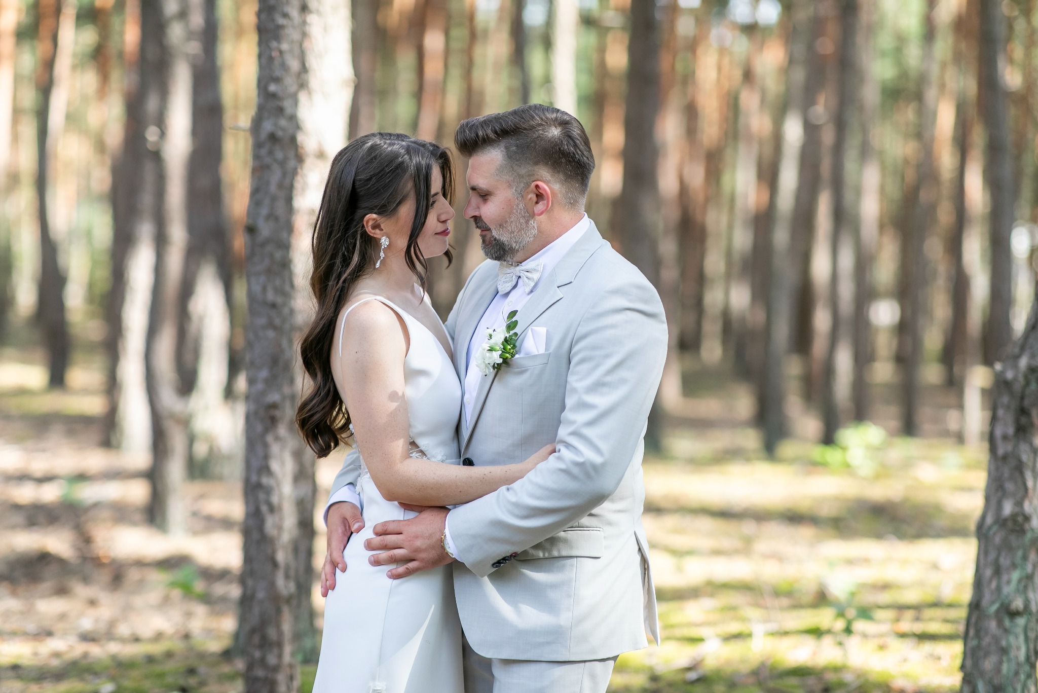 Para młoda w jasnych strojach, obejmująca się czule w zalesionym plenerze. Delikatne światło podkreśla naturalność scenerii i intymność chwili.