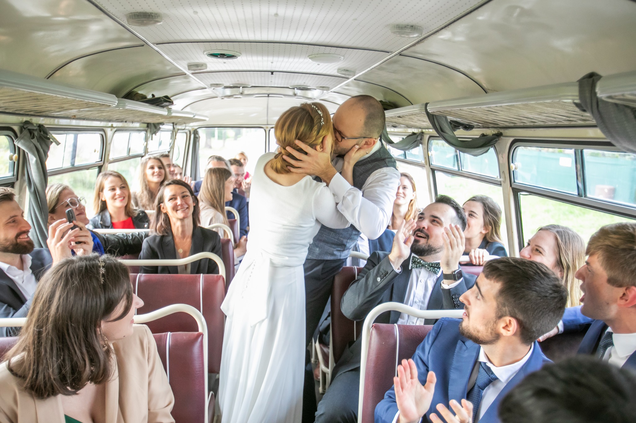Nowożeńcy całują się w autobusie pełnym gości weselnych, którzy im gratulują. Nietypowe ujęcie z wnętrza pojazdu, jasne oświetlenie, radosna atmosfera.
