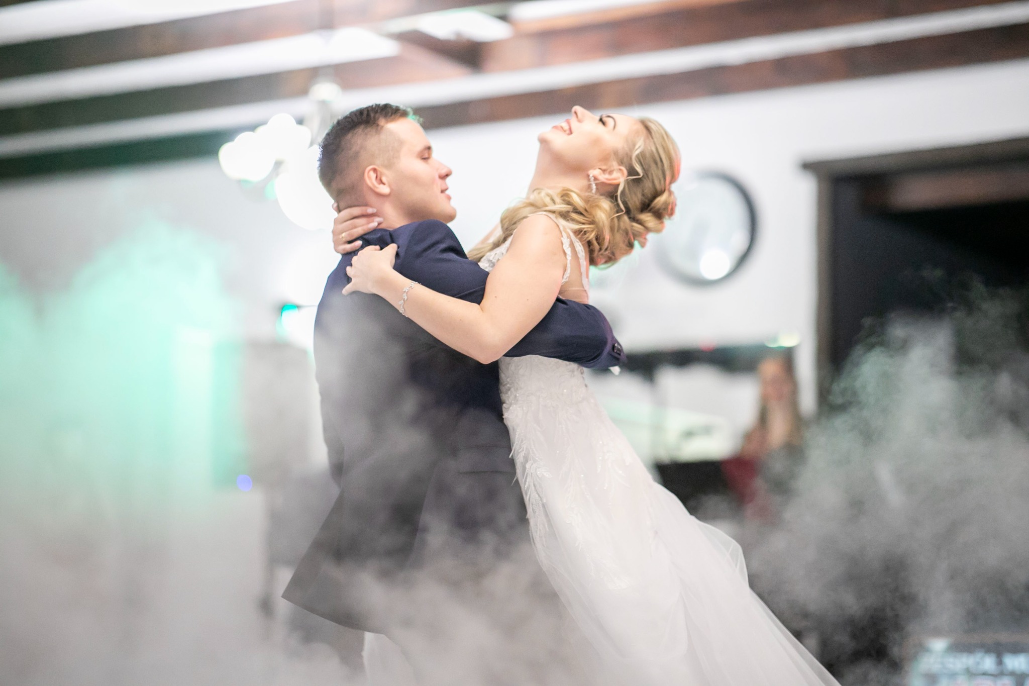 Młoda para w tańcu w obłokach dymu: panna młoda w białej sukni z odsłoniętymi ramionami i upiętymi włosami, pan młody w granatowym garniturze. Radosny nastrój.