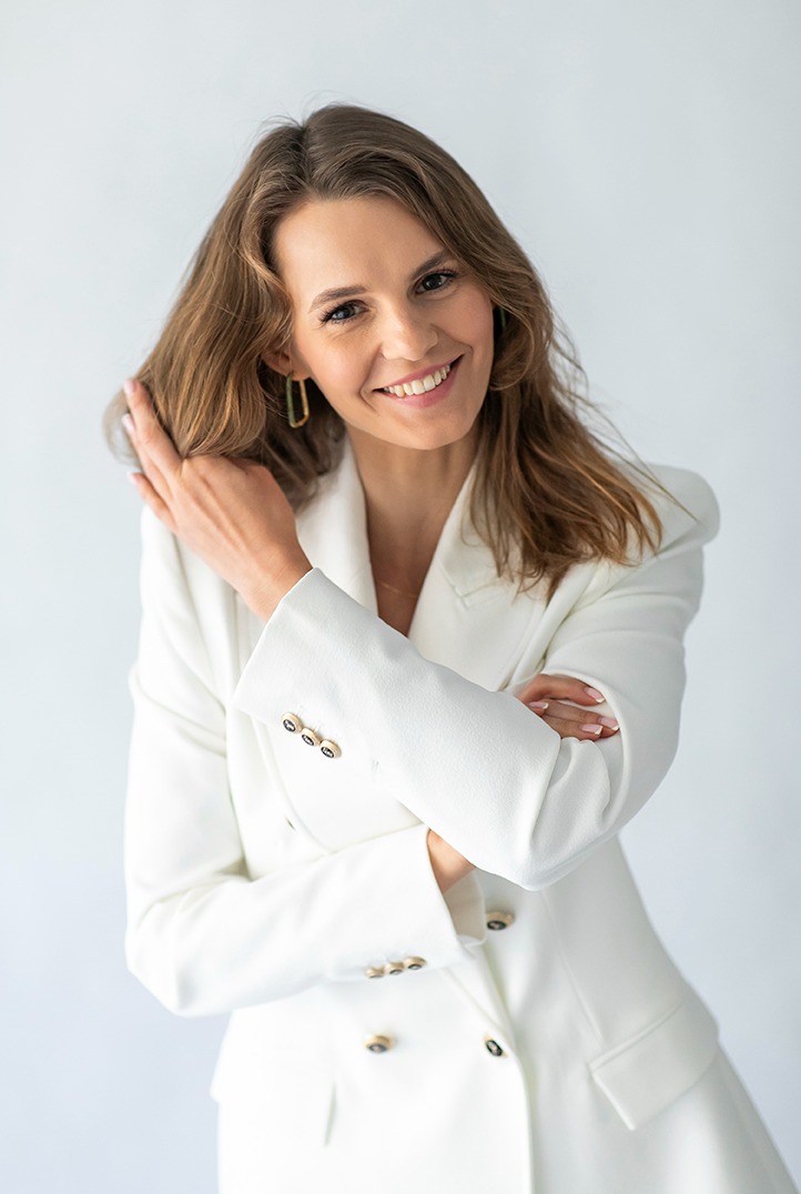 Uśmiechnięta brunetka w białym żakiecie, pozująca na jasnym tle. Kobieta dotyka włosów, ma złote kolczyki i skrzyżowane ręce. Portret biznesowy.