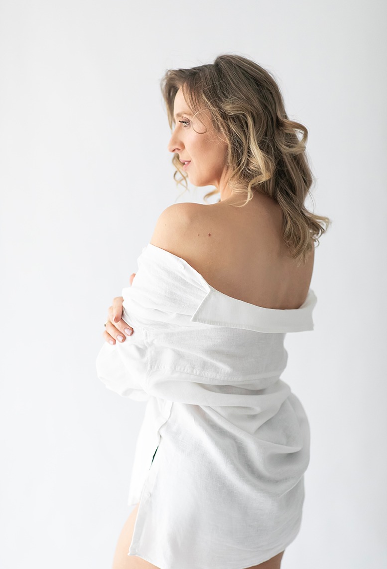 Portret kobiety z profilu, odwróconej tyłem, w białej koszuli, na jasnym tle. Widoczne delikatne fale we włosach i subtelny makijaż.