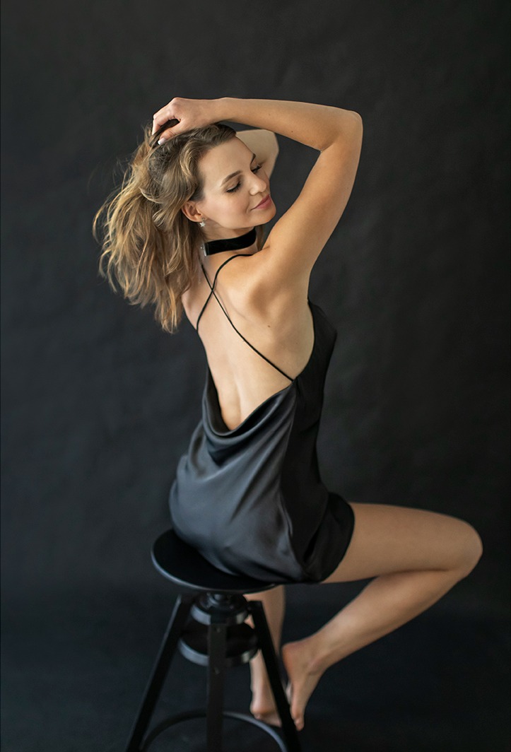 Kobieta w czarnej sukience, siedząca na stołku z ręką we włosach, patrząca w dół, z delikatnym uśmiechem, na tle ciemnego tła.