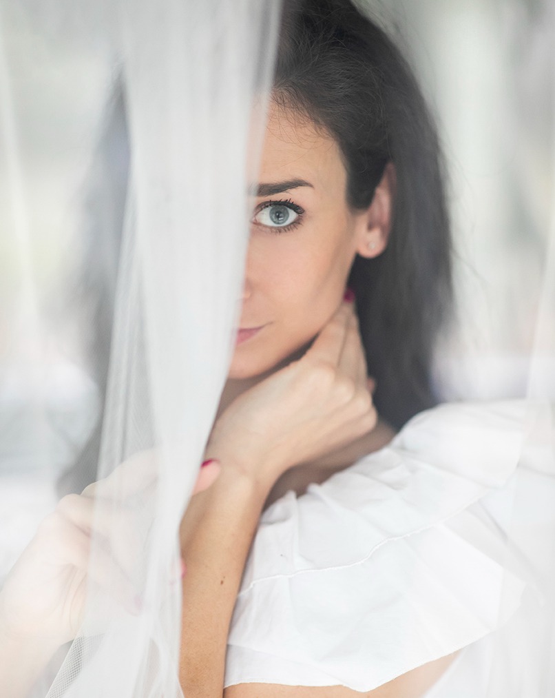 Portret kobiety o niebieskich oczach, częściowo zasłoniętej zwiewną tkaniną, z delikatnym makijażem i białej bluzce z falbanami. Subtelne światło.