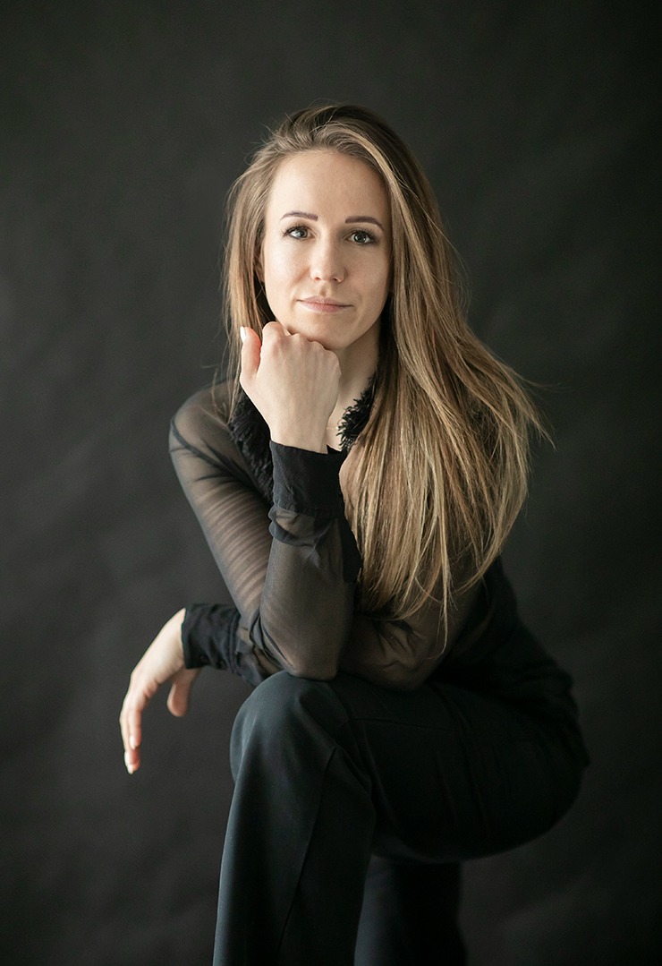 Portret kobiety w czarnej, prześwitującej bluzce, opierającej podbródek o dłoń, z długimi, prostymi włosami na ciemnym tle. Delikatny uśmiech, stonowana kolorystyka.