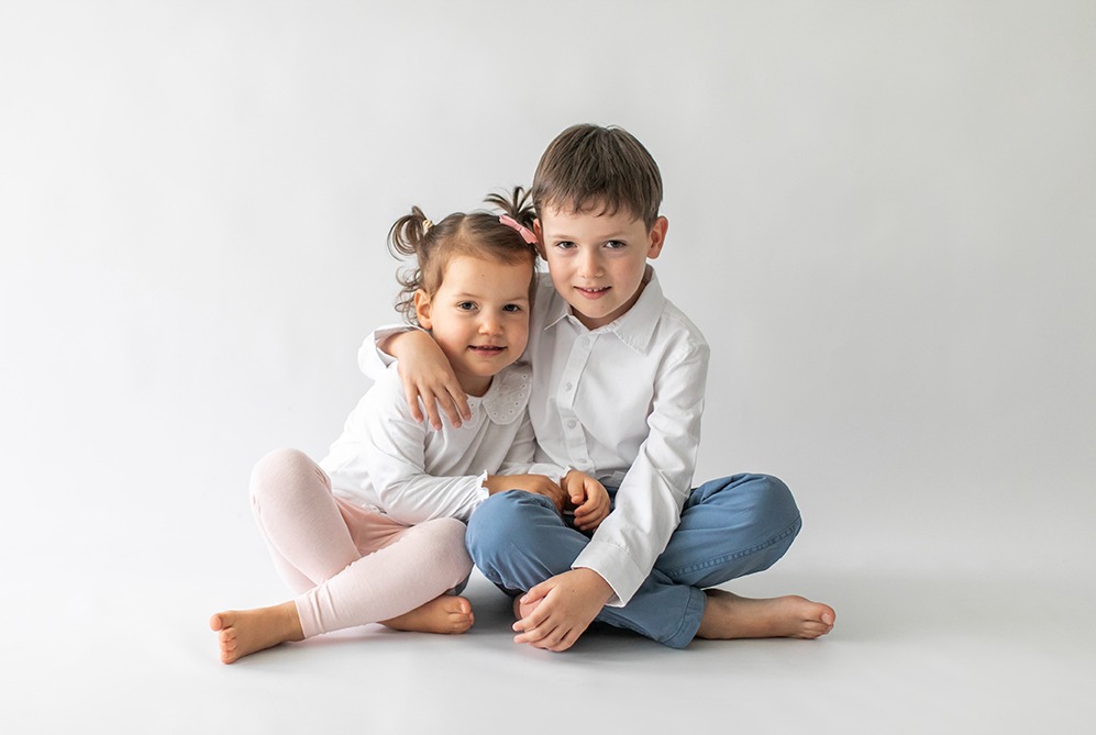 Uśmiechnięte rodzeństwo: dziewczynka z kokardą i chłopiec w białej koszuli, siedzą na podłodze w jasnym studiu, obejmując się, patrzą w obiektyw.