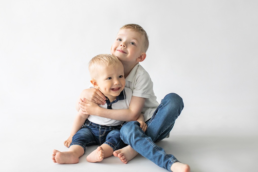Portret dwóch uśmiechniętych braci na jasnym tle: starszy obejmuje młodszego. Chłopcy ubrani w koszulki polo i dżinsy. Radosna, rodzinna atmosfera.
