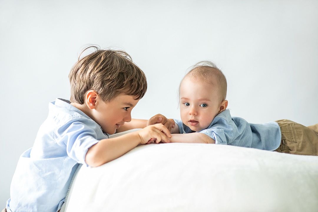 Jasne, studyjne ujęcie dwóch chłopców w błękitnych koszulach leżących na białej poduszce. Starszy brat z brązowymi włosami patrzy na młodszego, który spogląda w obiektyw.
