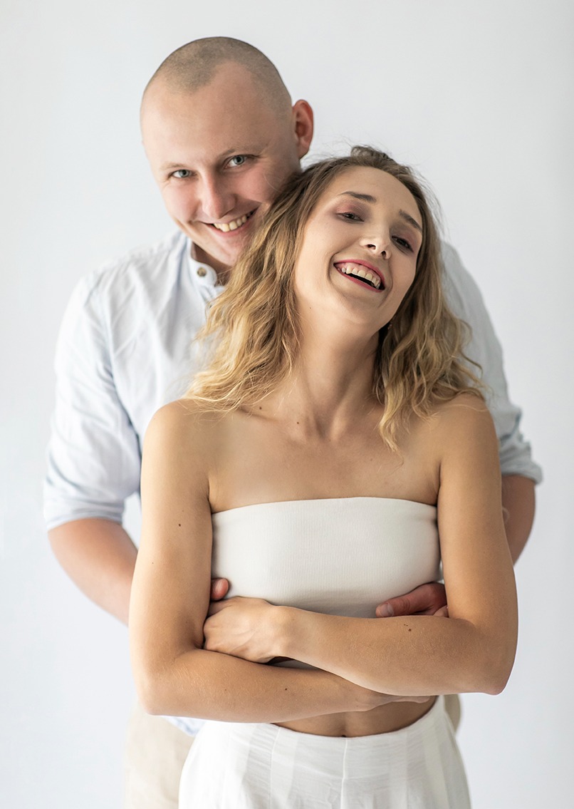 Uśmiechnięta para w jasnej tonacji: kobieta w białym topie bez ramiączek, obejmowana przez mężczyznę w błękitnej koszuli. Zdjęcie studyjne na białym tle.