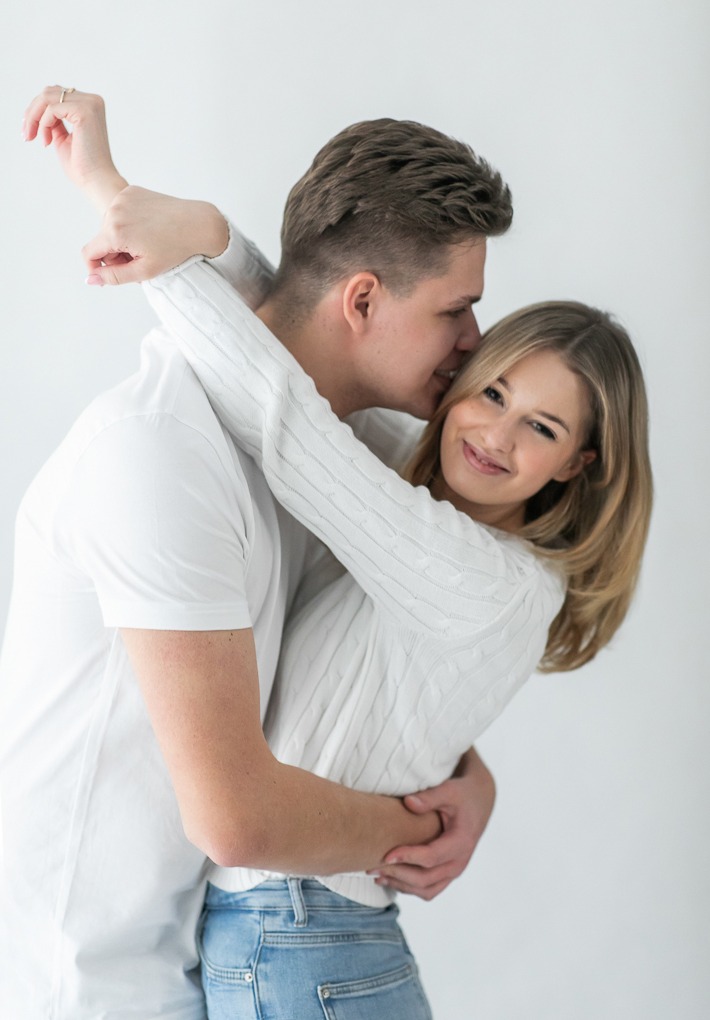 Uśmiechnięta blondynka w objęciach partnera, subtelny pierścionek na palcu, jasne tło, lekki uśmiech, bliskość, swobodna poza, naturalne światło.